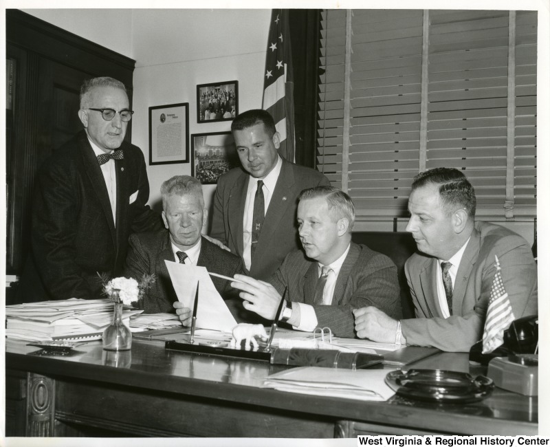 Congressman Arch Moore, Jr. sitting at his desk going over a document with four other unidentified individuals.