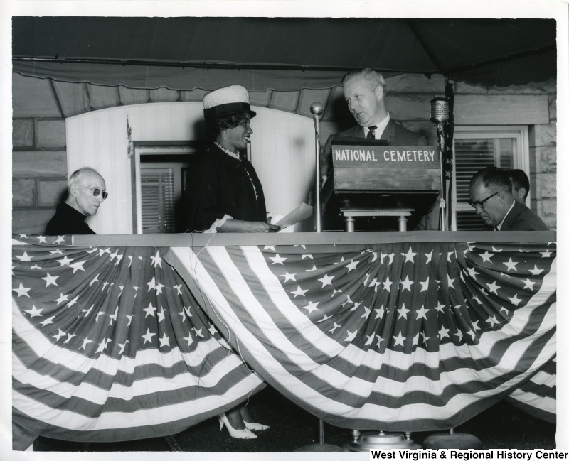 Congressman Arch A. Moore, Jr. talking with an unidentified African American woman while standing at a podium. The podium has a sign that reads, "National Cemetery."  Two unidentified men are sitting on both sides of Moore.