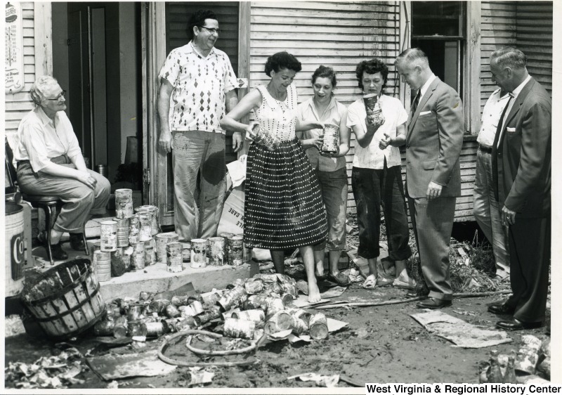 Three unidentified women are showing Congressman Arch A. Moore, Jr.damaged canned tomato juice. On the ground around them are stacks and piles of other canned goods.  An unidentified woman is sitting on the left side of the photo and three other unidentified men are present.