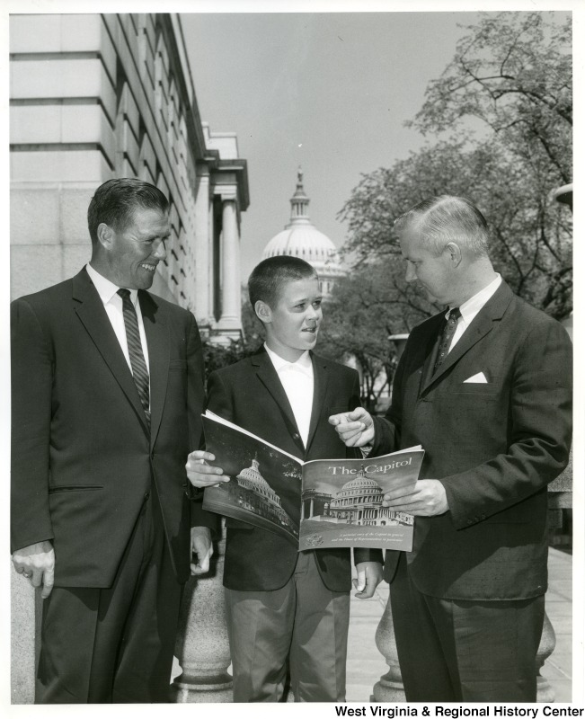 Congressman Arch A. Moore, Jr. standing with a man identified only as Baumgardner and his son.  The son is holding a copy of "The Capitol: A pictorial story of the Capitol in general and the House of Representatives in particular."