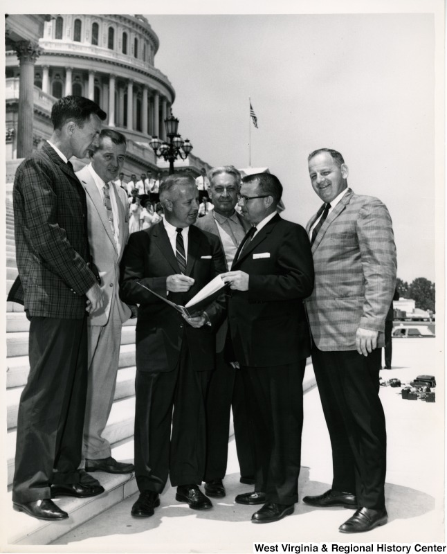 Congressman Arch A. Moore, Jr. showing a book to a group of unidentified men. They are standing on the steps of the Capitol Building.