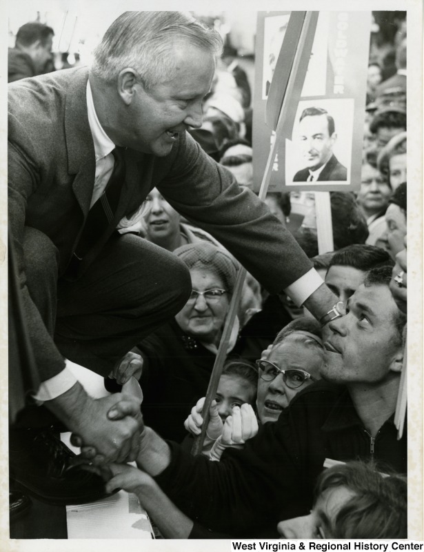 Congressman Arch A. Moore, Jr. crouching down to shake the hand of an unidentified man in the crowd during a Barry Goldwater rally in Wheeling.