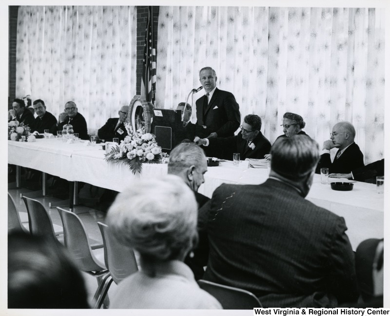 Congressman Arch A. Moore, Jr. standing at a tabletop podium speaking.  Seated at the table is a group of unidentified men. The photo is taken at an angle in the audience.