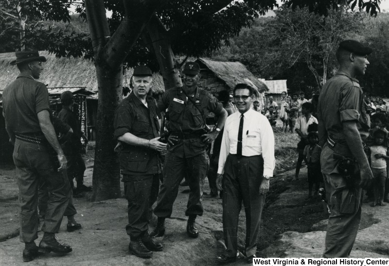 Congressman Arch A. Moore, Jr. talking to an unidentified man in a white dress shirt and a man in uniform. They are standing in a village or refugee camp in Vietnam.