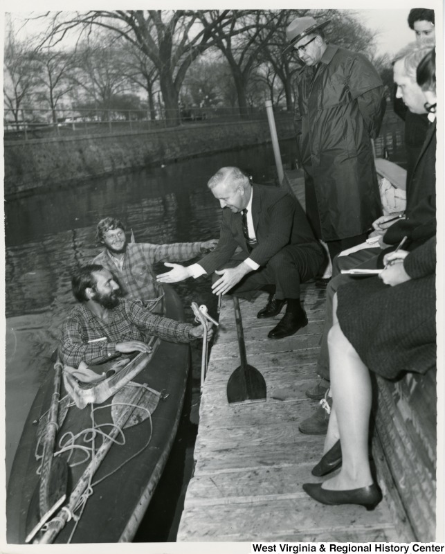 Congressman Arch A. Moore, Jr. is crouched down talking to two men in a kayak. Moore is holding out his hand. An officer is standing beside Congressman Moore.