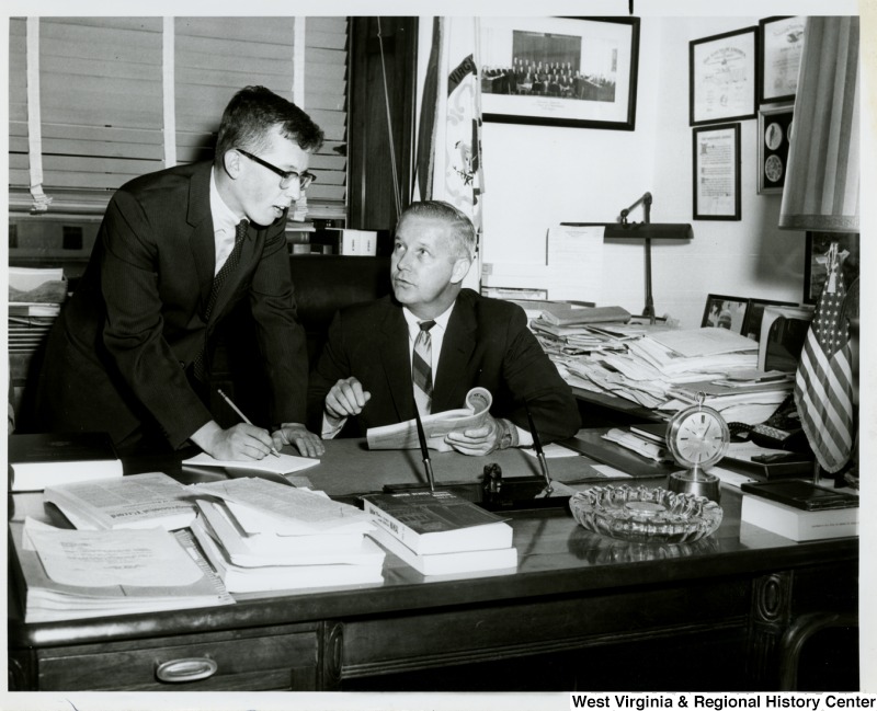 Congressman Arch A. Moore, Jr. seated at his desk talking to an unidentified man. The man appears to be taking notes.