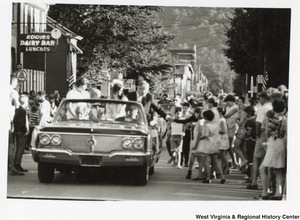 Congressman Arch Moore and his wife, Shelley, riding in the back of a convertible during a parade. He is shaking hands with a bystander.