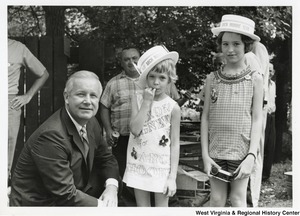 Congressman Arch Moore crouched beside two little girls who are wearing Team Arch Moore hats, and Moore and Nixon campaign buttons. The little girl closest to Arch is wearing a dress that says Nixon/Agnew for Arch Moore.