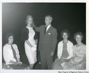 Congressman Arch Moore, Jr. (center) standing next to an unidentified woman wearing a tiara. Two unidentified women are sitting on the right and one on the left.