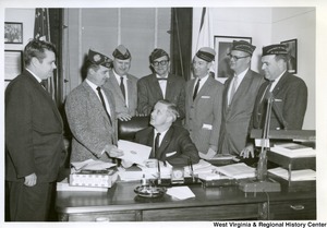 Congressman Arch A. Moore, Jr. (seated) being handed a document from a member of the Veterans of Foreign Wars.  Six other unidentified members are standing around the desk watching.