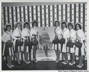 Eight unidentified women wearing Re-Elect Congressman Arch A. Moore, Jr. sashes and holding baskets. In the center is a poster of Moore walking away from the Capitol.
