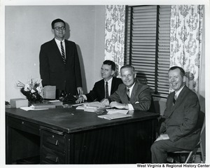 Congressman Arch A. Moore, Jr. seated at a desk with two unidentified men. Another unidentified man is standing on the far side (left) against the wall.