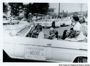 Congressman Arch A. Moore, Jr. and his family in the back of a convertible during a parade. Mrs. Shelley Moore and their daughter Lucy, are shown. Mrs. Moore is holding a bouquet of flowers. Lucy is leaning against the back of the driver seat. A sign on the side of the vehicle reads "Congressman Arch Moore, Jr. and family."