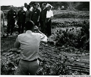 A photographer is taking a photograph of a group of men, including Congressman Arch A. Moore, Jr., in a muddy field in Vietnam.