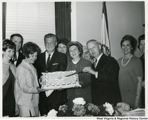 Congressman Arch A. Moore, Jr. pointing, with a knife, to his birthday cake, which is being held up by Mrs. Shelley Moore and two others. They are surrounded by people.