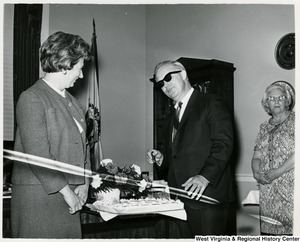 Congressman Arch A. Moore, Jr. talking to his wife, Shelley, at his birthday party. He appears ready to cut the cake. Moore is also wearing a pair of sunglasses.