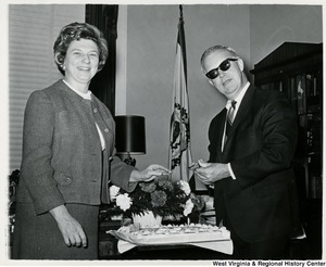 Congressman Arch A. Moore, Jr. and his wife, Shelley, at his birthday party. Moore is holding a knife and appears to be getting ready to cut the cake. Moore is also wearing sunglasses.