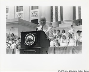 Governor Arch Moore Jr speaking at a podium that has the Seal of West Virginia on it. People are seated behind the Governor.