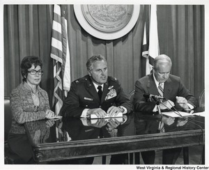 Governor Arch Moore (first on right) sitting with an unidentified man in uniform and an unidentified woman at what appears to be a press conference or hearing. The great seal of West Virginia is behind them.