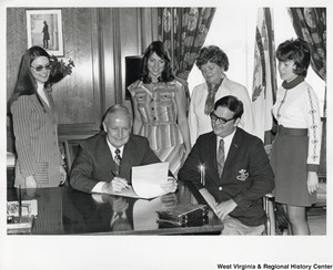 Governor Arch Moore (seated, first on left) signing a document while an unidentified man and four unidentified women watch.