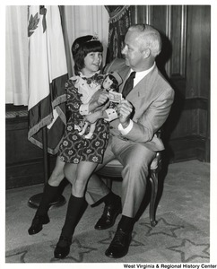 Governor Arch Moore with an unidentified little girl sitting on his lap holding a doll. She and the Governor are holding onto a folded one-dollar bill.