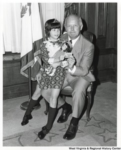 Governor Arch Moore with an unidentified little girl sitting on his lap holding a doll. She and the Governor are holding onto a folded one-dollar bill.