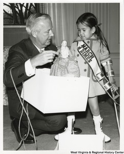 Governor Arch Moore helping a little girl with crutches open a present of a doll. The girl is wearing a March of Dimes sash.