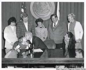 Governor Arch Moore kissing a little kid on the cheek. An unidentified woman is sitting to the right of Governor Moore. Three unidentified women and two men are standing behind the governor.
