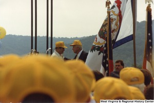 Governor Arch Moore speaking to an unidentified man on a stage for the Moundsville Bridge Dedication. Moore, the unidentified man, and the audience are wearing yellow hats.