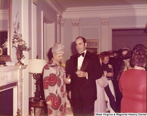 An unidentified man and woman holding drinks at a reception. There are other attendees in the background socializing.
