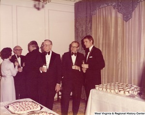 Five men and one woman are mingling next to two table of food and drink.