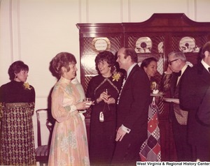 Two women and a man having a conversation at a reception. They are surrounded by other guests.