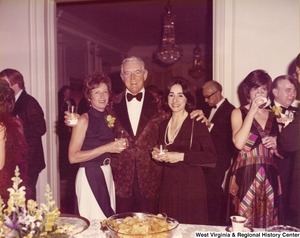 An unidentified man has his arms around two women at a reception. They are standing behind a table with food and are surrounded by other guest.