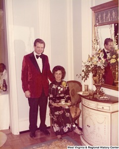 An unidentified man and woman at a reception. The woman is seated on a chair holding a plate and a drink.