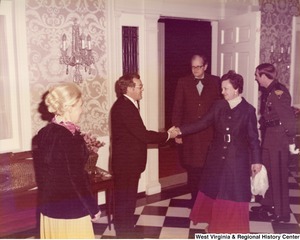 An unidentified man shakes a womans hand as she comes through the door. An unidentified man is halfway through the door. An unidentified woman (back towards the camera) is waiting to greet the man and woman. The door is being held open by an unidentified state trooper.