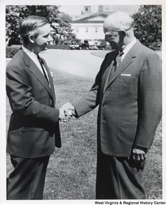 Congressman Arch Moore shaking the hand of an unidentified man. They are standing outside. A house and two cars can be seen in the background.