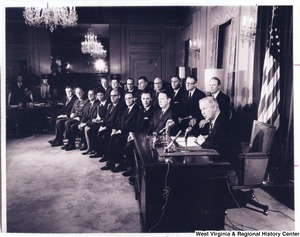 Governor Arch Moore speaking to the press. The Governor is seated at a desk in front of the American Flag. To the left of the Governor is two rows of people sitting for his announcement.