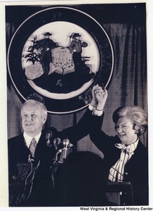 Governor Arch Moore and his wife, Shelley, holding their joined hands into the air. They are standing at a podium speaking and the Seal of West Virginia is behind them.