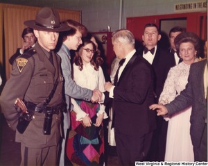 Governor Arch Moore shaking the hand of an unidentified man as they come through a doorway of a reception. Shelley Moore is standing to the right of Arch. They are surrounded by unidentified men and an unidentified State Police officer.