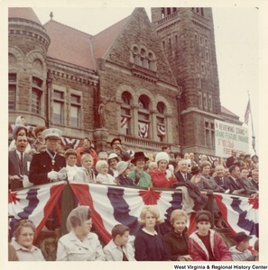 Governor Arch Moore, Jr. (fourth from the right) sitting with his wife Shelly in raised seating watching the 31st Mountain State Forest Festival Parade. The seating is in front of the Randolph County Courthouse.
