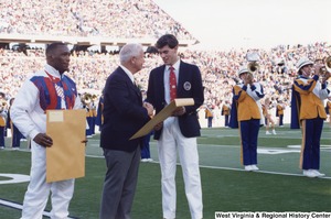 Governor Arch Moore (center) shaking the hand of Olympian Web Wright (right) and handing him a large manila envelope on the West Virginia University football field. Olympian Nate Carr (left) is standing beside Governor Moore holding a large manila envelope. The West Virginia University marching band is playing in the background.