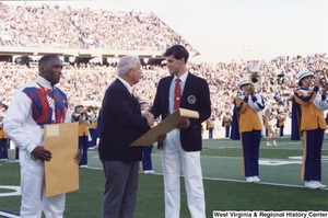 Governor Arch Moore (center) shaking the hand of Olympian Web Wright (right) and handing him a large manila envelope on the West Virginia University football field. Olympian Nate Carr (left) is standing beside Governor Moore holding a large manila envelope. The West Virginia University marching band is playing in the background.