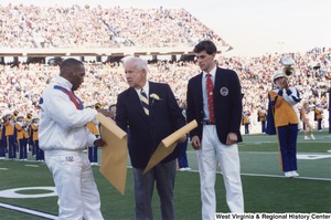 Governor Arch Moore (center) handing a large manila envelope to Olympic wrestler Nate Carr. Olympic shooter Webster Wright is standing to the right of Governor Moore. The West Virginia University marching band is playing in the background.