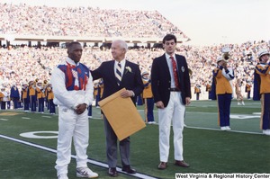 Governor Arch Moore standing between Olympic athletes Nate Carr (left) and Webster Wright (right). Governor Moore is holding a large manilla envelope. The West Virginia University marching band is playing in the background and the stands are filled with people.