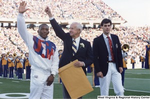 Governor Arch Moore standing between Olympic athletes Nate Carr (left) and Webster Wright (right). Governor Moore has one arm raised and is holding a large manilla envelope in the other hand. The West Virginia University marching band is playing in the background and the stands are filled with people.