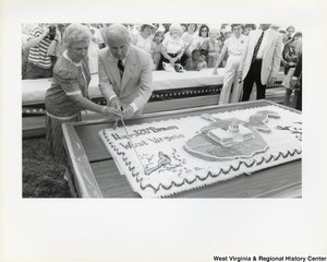Governor Arch Moore and his wife Shelley Moore cutting a large birthday cake that says Happy 125th Birthday West Virginia and features the state of West Virginia, the Capitol building, and a cardinal.