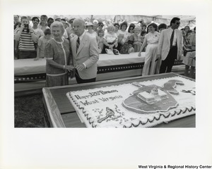 Governor Arch Moore and his wife Shelley Moore standing behind a large birthday cake that says Happy 125th Birthday West Virginia and features the state of West Virginia, the Capitol building, and a cardinal.