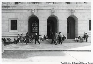 Governor Arch Moore shaking the hand of an unidentified man. The photo is taken from a distance. Four unidentified men are standing a short distance behind the Governor. Groups of unidentified people are sitting on the steps watching the Governor.