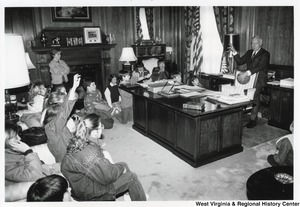Governor Arch Moore standing behind his desk holding a basketball, is talking to a group of kids who are seated around his office.