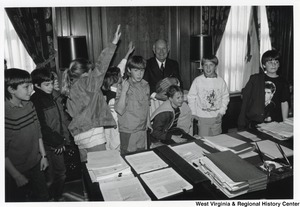 Governor Arch Moore standing behind his desk with a group of kids. The Governor is holding a basketball and leaning on the back of his chair. A unidentified kid is sitting in his chair.